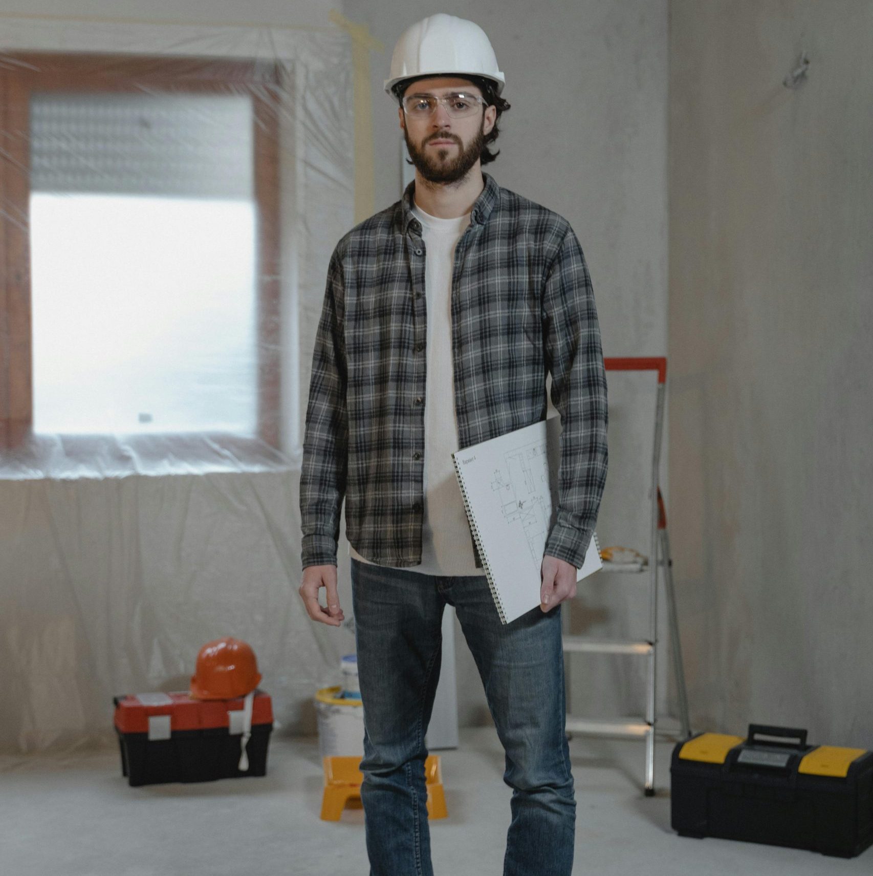 Homme debout dans un batiment en construction avec casque de chantier et un dossier dans la main gauche